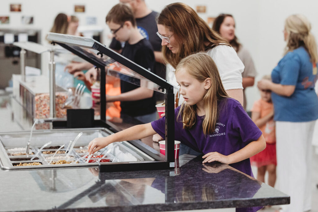 A woman and a girl, both smiling, select toppings at a dessert bar while others are in the background enjoying treats.