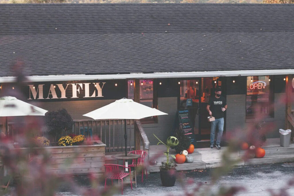 A cozy cafe named "Mayfly" with outdoor seating, pumpkins, and festive lights, set against an autumn backdrop.
