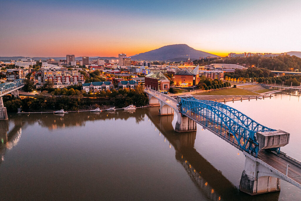 A scenic view of a city at sunset, featuring a blue bridge over a river and a mountain in the background.