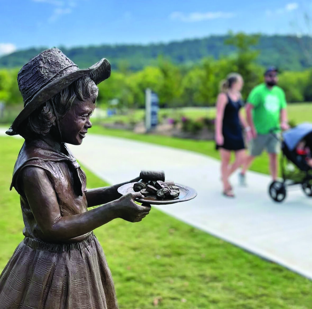 Bronze "Little Debbie" Sculpture with couple walking on sidewalk in the background pushing a stroller